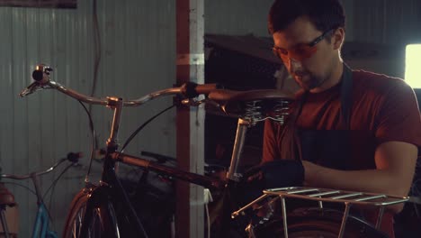 theme-small-business-bike-repair.-A-young-Caucasian-brunette-man-wearing-safety-goggles,-gloves-and-an-apron-uses-a-hand-tool-to-repair-and-adjust-the-bike-in-the-workshop-garage