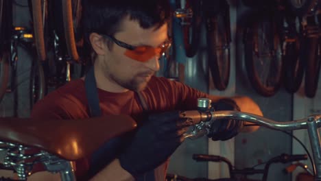 theme-small-business-bike-repair.-A-young-Caucasian-brunette-man-wearing-safety-goggles,-gloves-and-an-apron-uses-a-hand-tool-to-repair-and-adjust-the-bike-in-the-workshop-garage