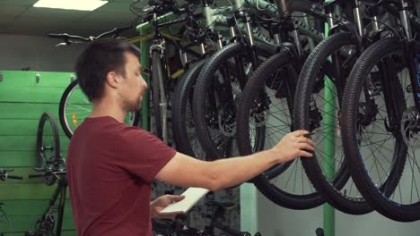 theme-of-small-business-selling-bicycles.-Young-Caucasian-male-brunette-small-business-owner,-store-manager-uses-notepad-and-pen-makes-notes,-checklist-at-bicycle-store