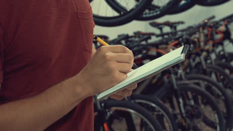 theme-of-small-business-selling-bicycles.-Young-Caucasian-male-brunette-small-business-owner,-store-manager-uses-notepad-and-pen-makes-notes,-checklist-at-bicycle-store