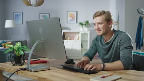 Porträt-von-hübscher-Mann-arbeiten-am-PC-sitzend-an-seinem-Schreibtisch.-Im-Hintergrund-stilvolle-gemütliche-Wohnzimmer.-Junge-freier-Mitarbeiter-arbeiten-von-zu-Hause-aus.