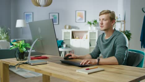 Portrait-of-the-Focused-Young-Man-Working-on-a-Personal-Computer-while-Sitting-at-His-Desk.-In-the-Background-Cozy-Living-Room.