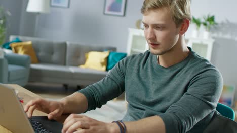 Gut-aussehend-lächelnde-jungen-Mann-mit-Laptop-beim-Sitzen-am-Schreibtisch-von-seiner-gemütlichen-Wohnzimmer.