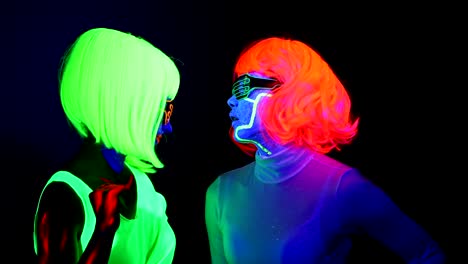 Women-with-UV-face-paint,-wig,-glowing-glasses,-glowing-clothing-dancing-together-slowly-in-front-of-camera,-Half-body-shot.-Caucasian-and-asian-woman.-.