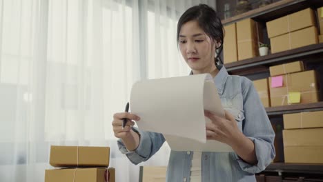 Beautiful-smart-Asian-young-entrepreneur-business-woman-owner-of-SME-checking-product-on-stock-and-write-on-clipboard-working-at-home.-Small-business-owner-at-home-office-concept.