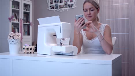 Young-woman-with-phone-sitting-at-the-table-next-to-sewing-machine