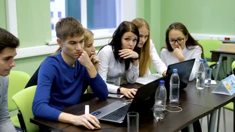 Team-of-young-managers-is-sitting-together-at-the-table-with-laptops-and-participating-in-competition-in-business-game-simulator.-Four-young-girls-and-two-boys