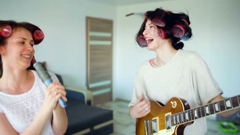 Two-funny-girls-singing-with-comb-and-playing-electric-guitar-dance,-sing-and-have-joy-at-home