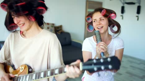 Dos-chicas-divertidas-cantando-con-el-peine-y-tocando-el-baile-de-la-guitarra-eléctrica,-cantan-y-tienen-alegría-en-casa