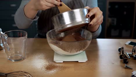 Close-up-view-of-young-woman-weighs-the-cocoa,-adds-to-flour.-Girl-using-kitchen-scales,-bowl-and-sieve-for-this