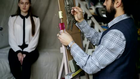 Sculptor-creating-sculpture-of-human's-face-on-canvas-while-young-woman-posing-him-in-art-studio