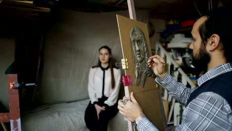 Bildhauer-Skulptur-des-menschlichen-Gesichts-auf-Leinwand-während-die-junge-Frau-posiert-ihn-im-Kunstatelier-erstellen