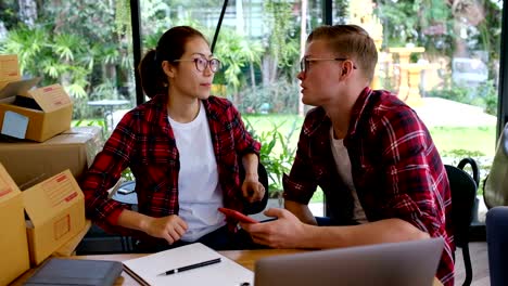 Inicio-pequeño-empresario-hablando-en-el-lugar-de-trabajo.-vendedor-freelance-cheque-orden-de-producto-con-smartphone-en-la-tienda.-Joven-pareja-mira-el-teléfono-móvil.-Online-venta,-comercio-electrónico,-envío-concepto