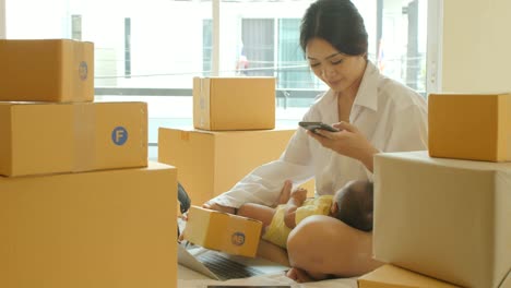 Young-asian-mother-holding-baby-and-checking-order-from-smartphone-for-customer-and-online-delivery-for-ready-packing-in-bedroom.
