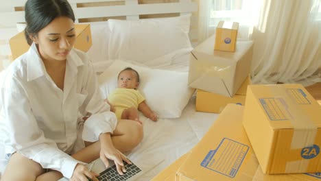 Young-asian-mother-and-her-newborn-child-checking-order-from-laptop-for-customer-and-online-delivery-for-ready-packing-in-bedroom.