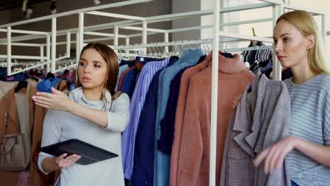 Young-businesswoman-is-checking-clothes-in-her-store-with-tablet-in-hands.-Her-employee-is-coming-and-asking-about-garment.-Employer-is-talking-to-her-and-gesturing.