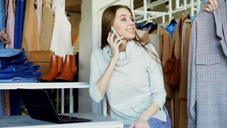 Mujer-propietaria-de-boutique-de-ropa-es-hablando-por-teléfono-móvil-y-escribiendo-en-la-laptop.-Su-asistente-se-le-acerca,-y-ella-está-dando-órdenes-de-manera-amistosa