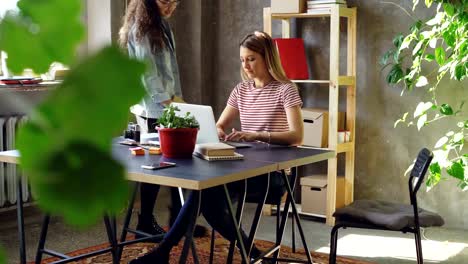 Joven-trabaja-con-ordenador-portátil-sentado-en-la-mesa-en-la-oficina.-Su-colega-está-viniendo,-las-mujeres-comienzan-mirando-la-pantalla-juntos-y-riendo.-Ambiente-informal.