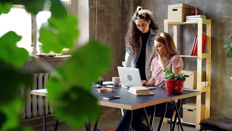Jóvenes-propietarios-de-pequeñas-empresas-están-trabajando-con-el-portátil-en-la-oficina-de-estilo-moderno-loft.-Rubia-está-sentada-y-escribiendo,-morena-es-permanente-y-sugiriendo-ideas.