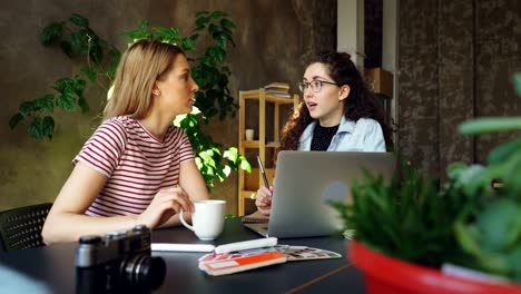 Studentinnen-sind-in-hellen-Raum-zusammensitzen-und-Geselligkeit.-Großer-Tisch-mit-Laptop,-Pflanze,-Teetasse-und-Markierungen-im-Vordergrund.-Ehrgeizige-Jugendkonzept.