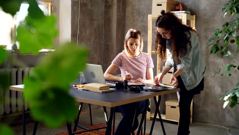 Jungen-Besitzer-des-Designstudios-arbeiten-zusammen-im-modernen-Büro.-Man-sitzt-und-schreiben,-andere-steht,-Kamera-halten-und-Verschieben-von-Fotos-auf-Tisch.