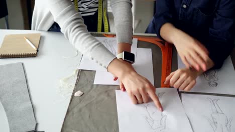 Close-up-shot-of-tailors'-hands-going-through-sketches-on-sewing-desk.-Women-are-discussing-clothing-designs.-Creative-dressmaker's-work-concept.