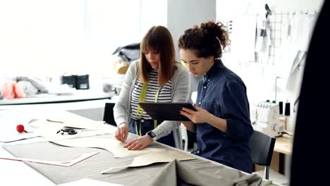 Attractive-clothes-designer-entrepreneurs-are-outlining-cutout-with-chalk,-working-with-tablet-and-talking.-Busy-day-at-successful-tailor's-shop-concept.