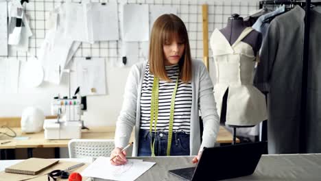 Beautiful-fashion-designer-is-drawing-clothing-sketches-for-her-new-clothes-collection-and-working-with-laptop.-Light-studio-with-garments-on-rails-and-drawings-in-background.