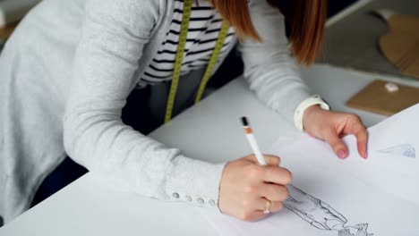 Close-tilt-down-shot-of-attractive-female-clothing-designer-drawing-sketch-with-pencil,-adding-shades-and-minor-details.-Woman-is-focused-on-her-work,-calm-and-thoughtful.