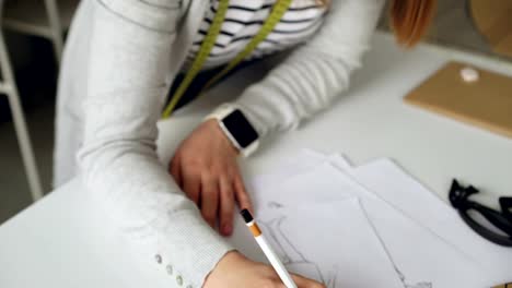 Tilt-up-of-Fashion-designer-is-drawing-lines-on-clothing-sketch-with-coffee,-tailor's-scissors,-ruler-and-pieces-of-paper-on-studio-table.-Attractive-girl-is-busy-and-concentrated-on-work.