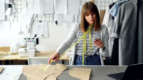 Young-seamstress-is-checking-clothing-paper-patterns-and-measuring-them-with-tape-measure-while-looking-at-smart-phone.-Nice-studio-with-garments-and-sewing-machine-in-background.