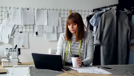 Junge-attraktive-Näherin-arbeitet-mit-Laptop-und-Kaffeetrinken-in-ihrem-Atelier.-Sie-hat-Ruhe-nach-der-Arbeit.-Entspannung-im-arbeitsplatzkonzept.