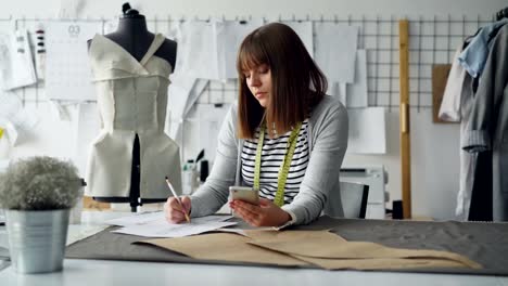 Creative-clothing-designer-is-looking-at-smartphone-and-drawing-sketch-while-working-in-modern-tailor-shop-at-sewing-table.-Woman-is-busy-and-involved-in-process.