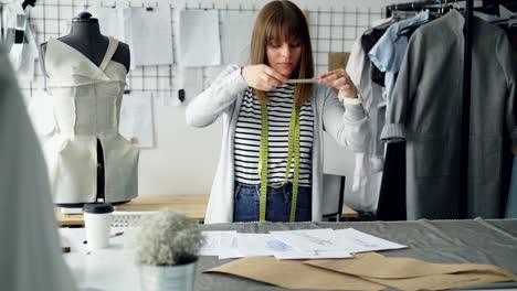 Ambitionierten-kreativen-weiblichen-Schneider-ist-Kleidungsstück-Skizzen-auf-Studio-Schreibtisch-platzieren-und-schießen-sie-mit-Smartphone.-Zeichnungen-der-neuen-Kollektion-Konzept-zu-präsentieren.