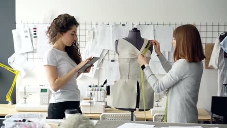 Professional-seamstress-is-measuring-patterns-on-mannequin-with-measure-tape-while-her-coworker-is-working-with-tablet-to-write-down-measurement-data.