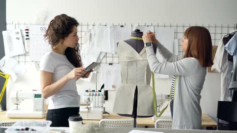 Clothing-designer-entrepreneurs-are-measuring-mannequin-parts-while-working-in-nice-modern-studio.-Young-attractive-woman-is-using-tablet-to-enter-measurement-results.