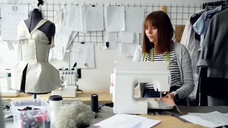 Professional-female-tailor-is-adjusting-sewing-machine-then-stitching-pieces-of-fabric.-Woman-is-working-in-light-tailoring-studio.-Sewing-items-and-mannequins-are-visible.