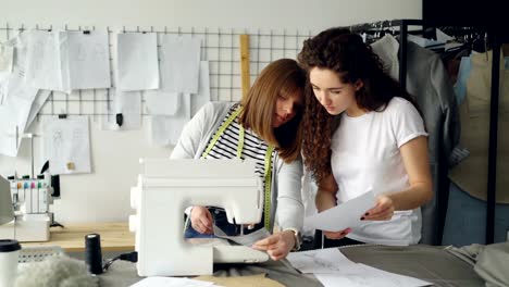 Attraktive-Schneiderinnen-sind-Blick-auf-Skizzen-und-arbeiten-mit-Nähmaschine,-dann-Stiche-Überprüfung-und-Anpassung-der-Ausrüstung.-Professionelles-Teamwork-Konzept.