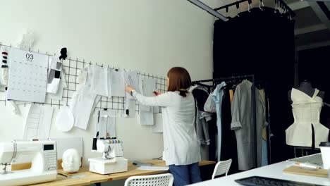 Female-clothing-designer-is-taking-sketches-from-studio-table-and-putting-them-on-wall-with-other-drawings-of-women's-garments.-Creative-thinking-concept.