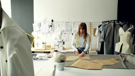 Young-attractive-female-designer-is-checking-her-paper-templates-with-measuring-tape.-Professional-creative-clothing-designing-concept.-Studio-is-light-and-modern.