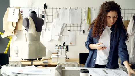 Clothing-designer-enterpreneur-is-shooting-flat-lay-of-garment-sketches-and-coffee-on-studio-desk-with-smart-phone.-Young-attractive-woman-is-moving-things-and-touching-screen.