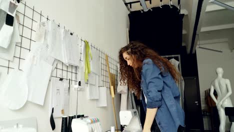 Niedrigen-Winkel-Blick-auf-junge-Mode-Designer-mit-Blick-auf-Skizzen-und-Zeichnungen-an-der-Wand-im-modernen-Loft-Stil-Studio-hängen.-Große-Sammlung-von-Abbildung-oben-Schneiderei-Schreibtisch.