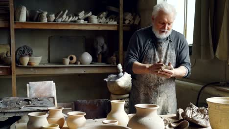 Professional-male-ceramist-is-kneading-clay,-forming-clay-ball-while-working-in-small-workshop-with-potter's-equipment,-tools-and-many-ceramic-figures.