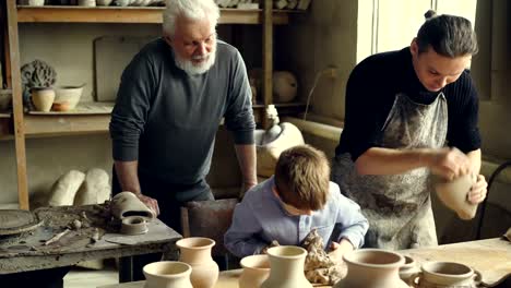 Professional-potter-is-kneading-clay-on-worktable-in-home-studio-while-his-son-is-helping-him-and-his-elderly-father-watching-them-from-behind.-Small-family-business-concept.