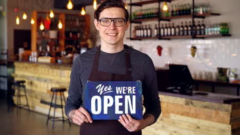 Porträt-von-fröhlich-hübscher-Junge-Kellner-Schürze-mit-\"Wir-sind-offen\"-Schild-im-Coffee-Shop-Eröffnung-stehen.-Start-Unternehmen-und-erfolgreiche-Menschen-Konzept.