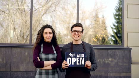 Happy-restaurant-owners-are-standing-outside-looking-at-windows-then-turning-to-camera-and-holding-open-sign-with-glad-proud-smile.-Successful-start-up-concept.