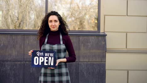 Portrait-of-attractive-confident-woman-in-apron-small-business-owner-holding-"yes-we-are-open"-sign-standing-outside-and-smiling-looking-at-camera.