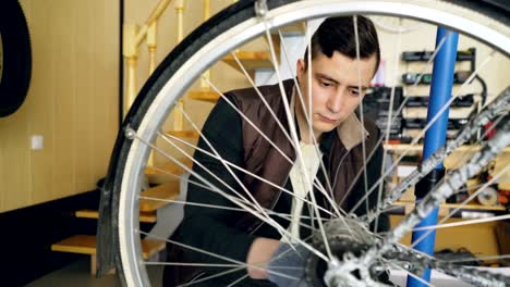 Junger-Mann-erfahrenen-Servicemann-ist-Fahrrad-Rad-mit-Schraubenschlüssel-und-Tools-Befestigung.-Kleine-gemütliche-Werkstatt-Interieur-mit-Holzwänden-und-Leiter,-Ersatzteile-und-Zubehör-sind-sichtbar.