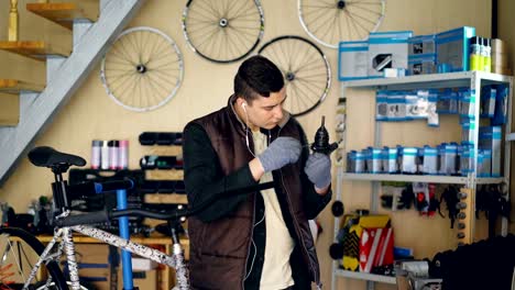 Concentrated-repairman-is-greasing-mechanism-while-repairing-bicycle-in-nice-workshop.-Young-man-in-warm-vest-and-protective-gloves-is-listening-to-music-.