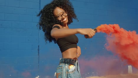 Beautiful-young-woman-holding-colorful-smoke-grenade-dancing-outside-against-blue-wall